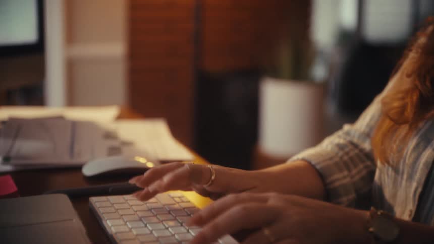 Slow motion close-up of a woman's hands typing on a computer keyboard in a dimly lit office