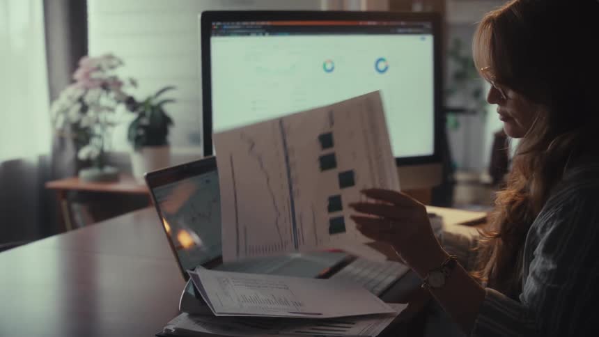 A focused woman in slow motion analyzes financial reports and data charts at her desk in a warmly lit home office