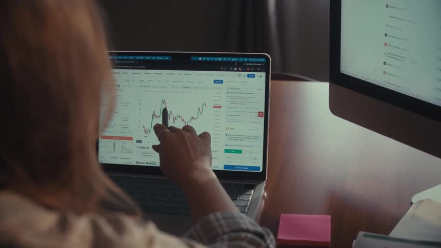 An over-the-shoulder shot of a financial analyst working at her desk, pointing at and analyzing a stock market graph on her laptop and second monitor