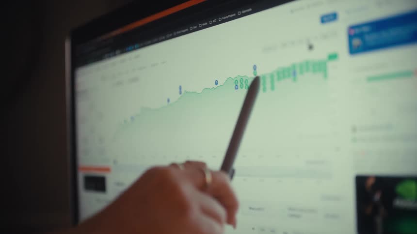 A financial analyst reviews market trends on a laptop and monitor, using a stylus to trace the growth on a stock chart