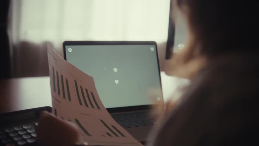 Over-the-shoulder view of a woman analyzing a financial report and pointing at a laptop with a green screen