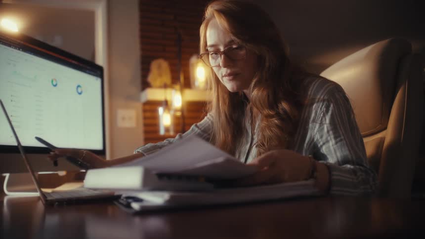 A diligent woman in glasses cross-references paperwork with data on her computer screen while working from home at night