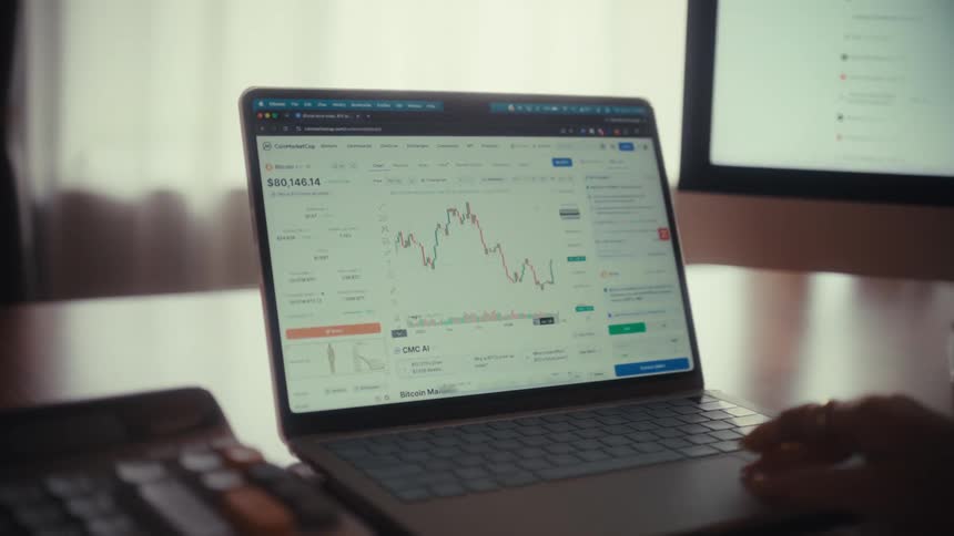 Close-up dolly out from a laptop screen showing a cryptocurrency price chart to reveal a person working and trading from their home office