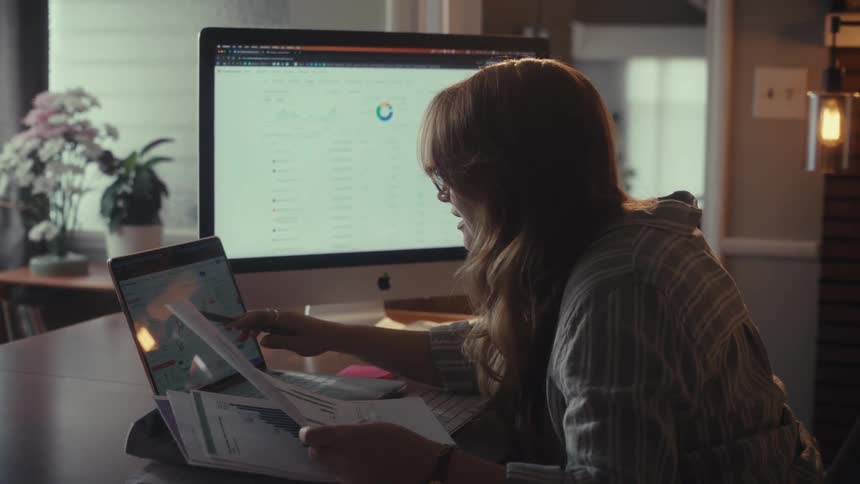 A professional woman analyzes financial data on multiple computer screens and paper reports in her home office