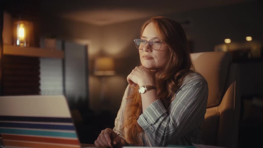 A contemplative woman with red hair and glasses works late from her cozy home office, pausing to think before continuing to type on her laptop