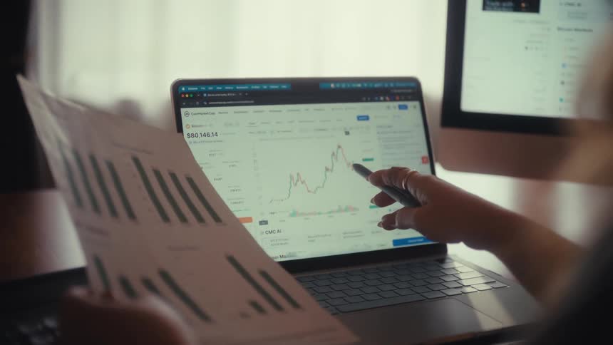 Over the shoulder shot of a financial analyst comparing printed bar charts with a live market trend graph on a laptop