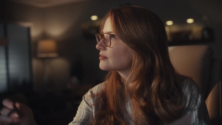 A young businesswoman with red hair and glasses analyzes financial charts and data on her computer while working late from home