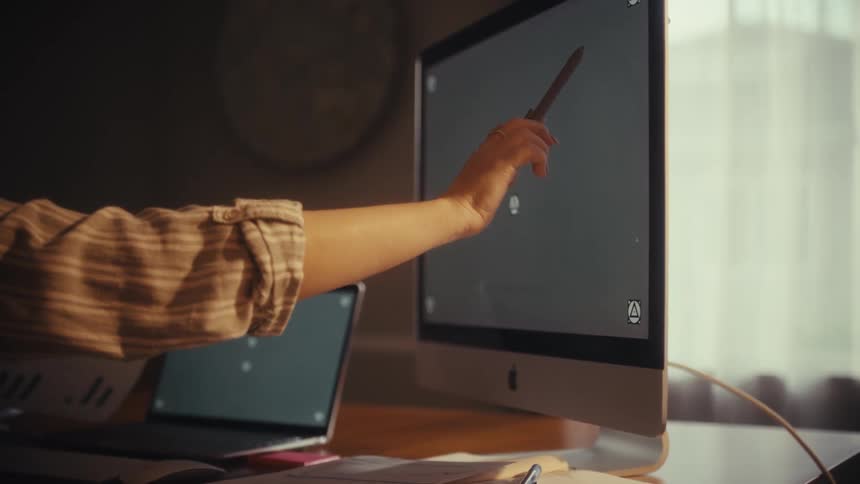 A woman in a home office setting analyzes charts while using a stylus to interact with her green-screen laptop and monitor