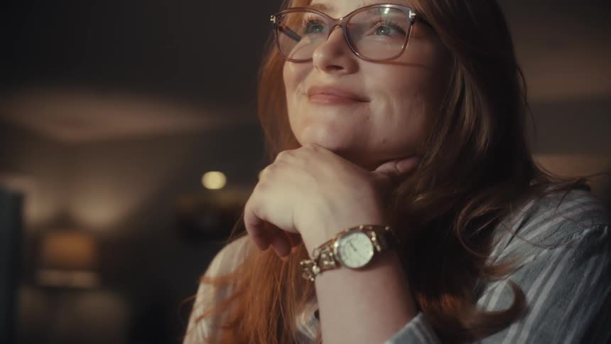 A happy young woman with red hair and glasses types on her keyboard and smiles with satisfaction while working late from home
