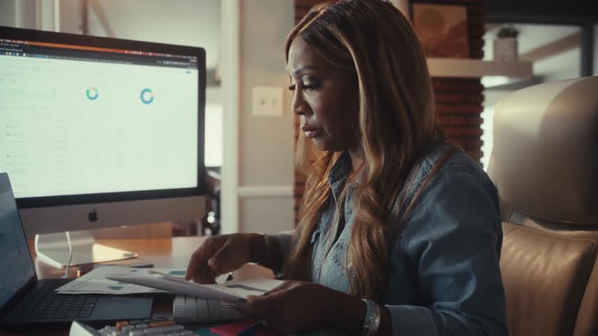 A professional woman meticulously analyzes financial reports and data on her computer in her home office in a cinematic slow-motion shot