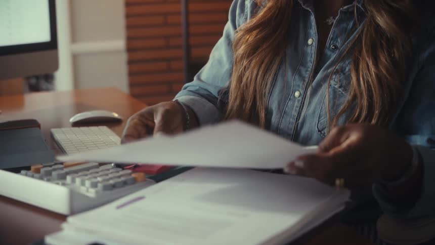 A woman in a denim shirt slowly flips through business reports with charts and graphs while seated at her home office desk