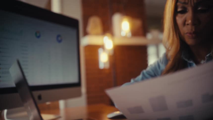 A concerned businesswoman analyzes financial data on her laptop and paper reports in her warmly lit home office