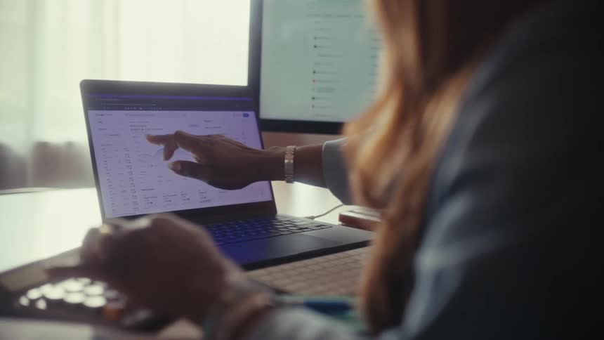 An over-the-shoulder shot of a businesswoman working at her desk, analyzing financial data and charts on her laptop with a dual-monitor setup