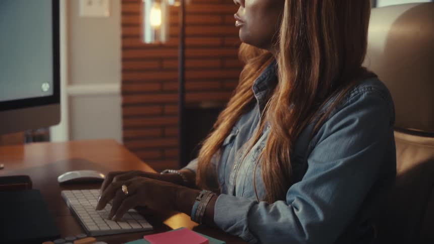 A businesswoman works at her desk, typing on the keyboard and pointing to the computer screen while reviewing work