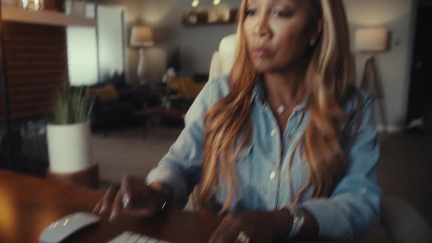 An African American woman types at her desk in a modern home office, then pauses to smile with satisfaction at her work