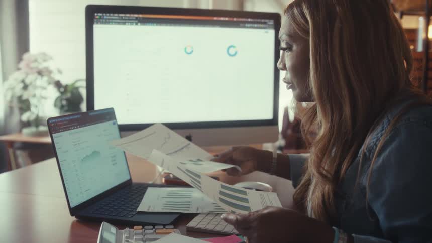 A focused woman reviews financial documents and compares data on her laptop and desktop computer while working from her cozy home office