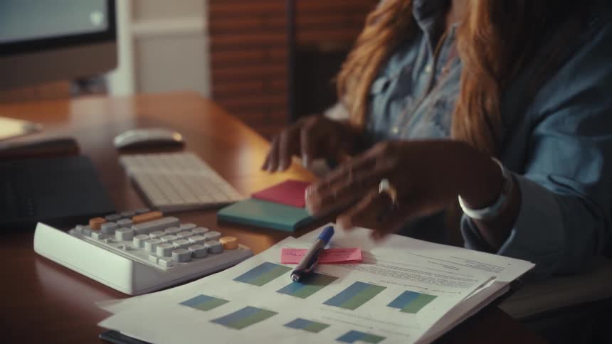 An African American woman working in her home office writes a sticky note reminder and types on her computer