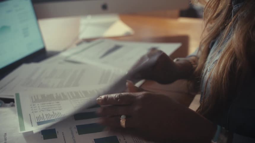 A concerned woman sorts through financial reports with charts and graphs, looking stressed while working at her desk