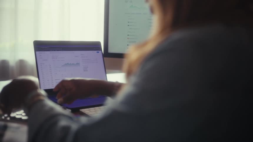 Over-the-shoulder shot of a financial analyst working on a laptop, checking data charts, and using a calculator at her desk