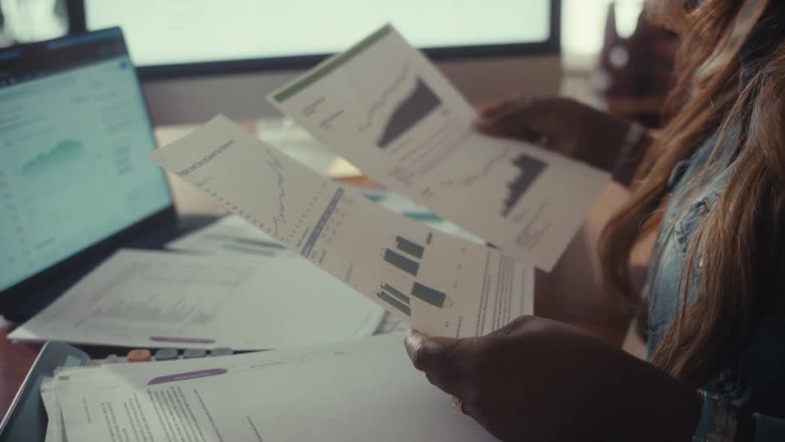 A businesswoman reviews financial documents and charts at her desk while working from home