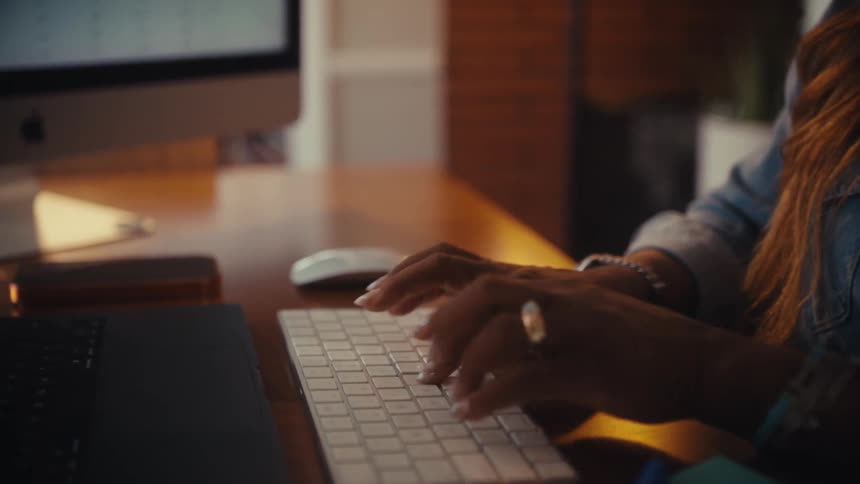 A close-up of a woman's hands typing on a wireless computer keyboard while working at her desk