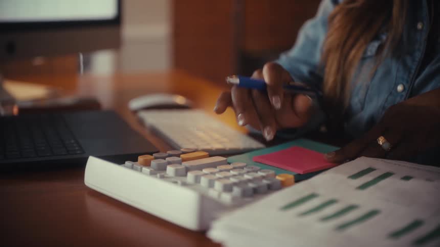 A businesswoman's hands use a calculator to analyze financial documents and charts at her office desk