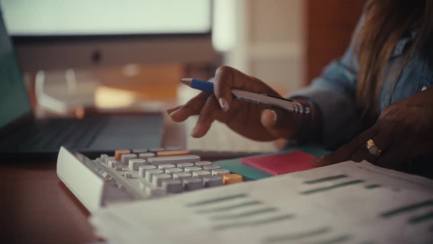 A business woman's hands use a calculator and take notes while reviewing financial reports at her desk