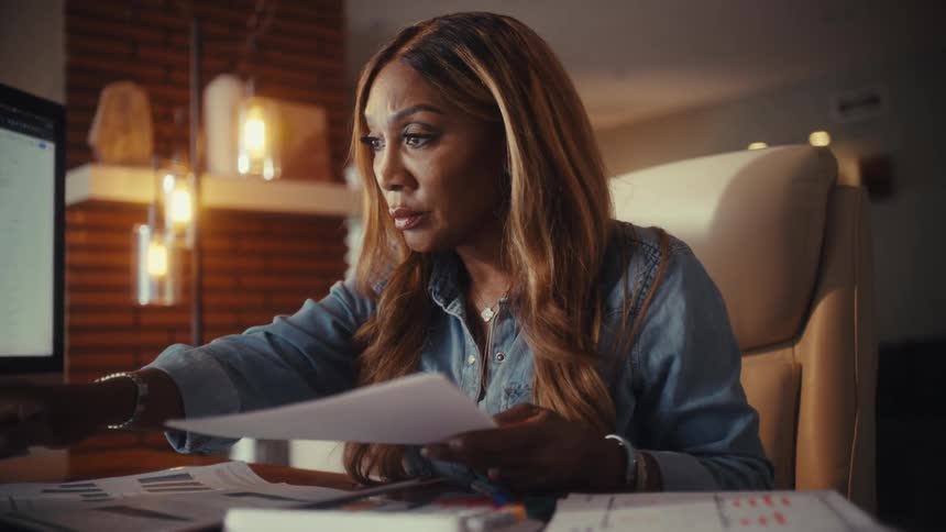 A businesswoman working from her home office at night analyzes financial reports on her computer, smiling with satisfaction at the results