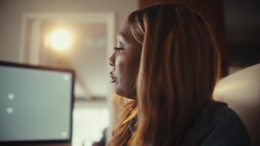 A focused woman works at her desk in a warmly lit home office, writing on paperwork with green screen computers in the background