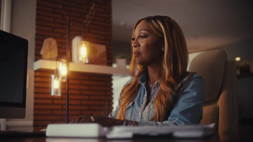 A stylish African American woman sits at her desk working diligently on her computer late at night in her modern home office