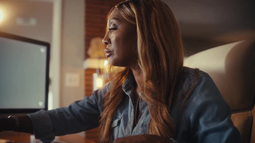 A focused Black woman works late at her home office desk, writing notes while referencing her computer screen