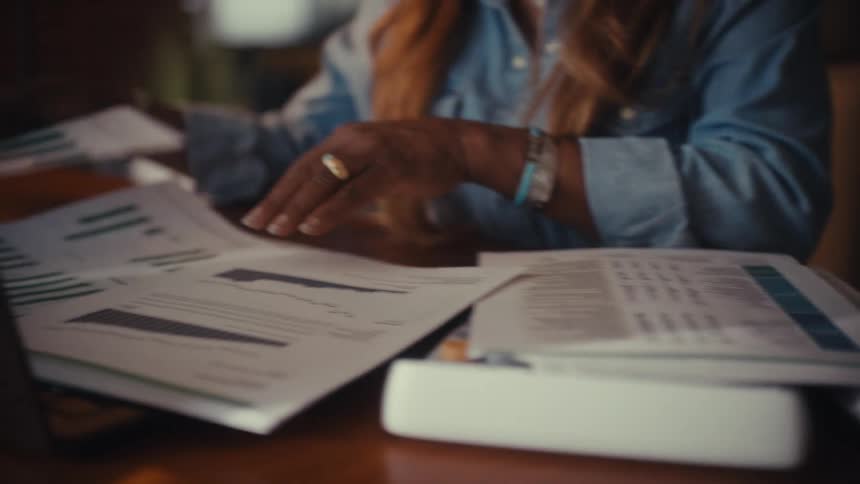 A businesswoman diligently reviews financial paperwork with graphs and charts at her desk in a warmly lit office