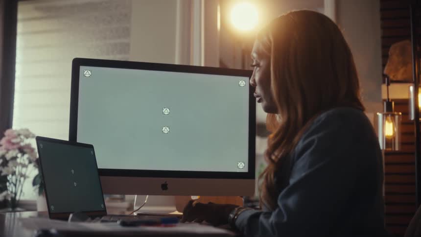 A focused woman works at her desk in a warmly lit home office on a computer and laptop with green screens