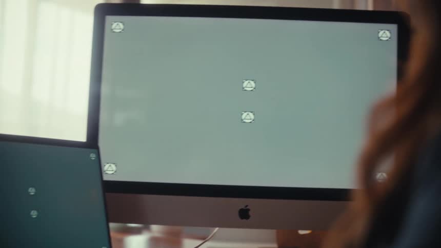 An over-the-shoulder shot of a woman working on a laptop and desktop computer with green screens, pointing to the monitor