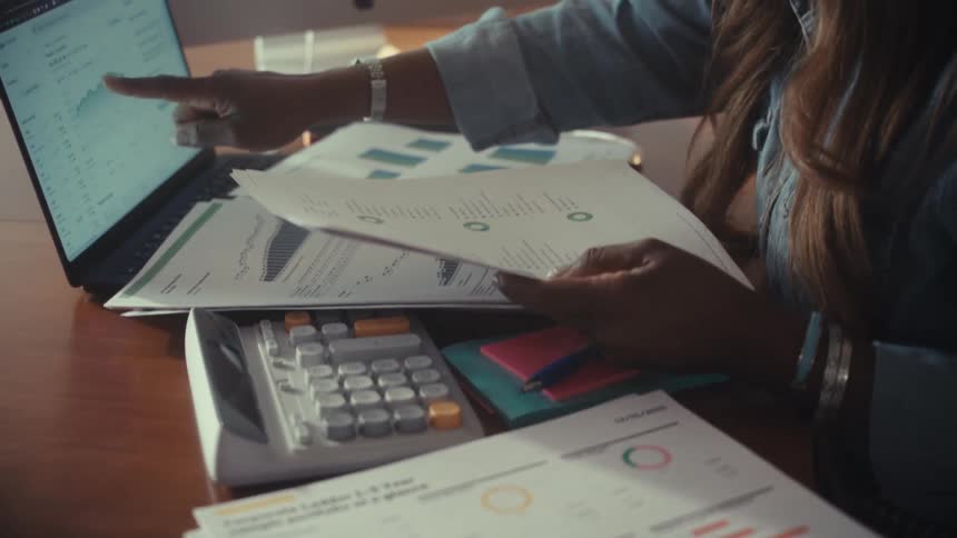 A Black woman reviews financial reports, comparing graphs on paper with data on her laptop at her desk