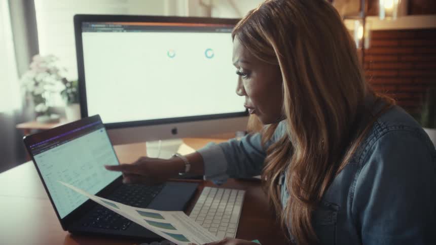 A concerned businesswoman analyzes financial data on her laptop and a printed report while working in her home office