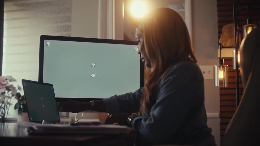A professional woman works from her stylish home office, multitasking between a desktop and laptop with green screens