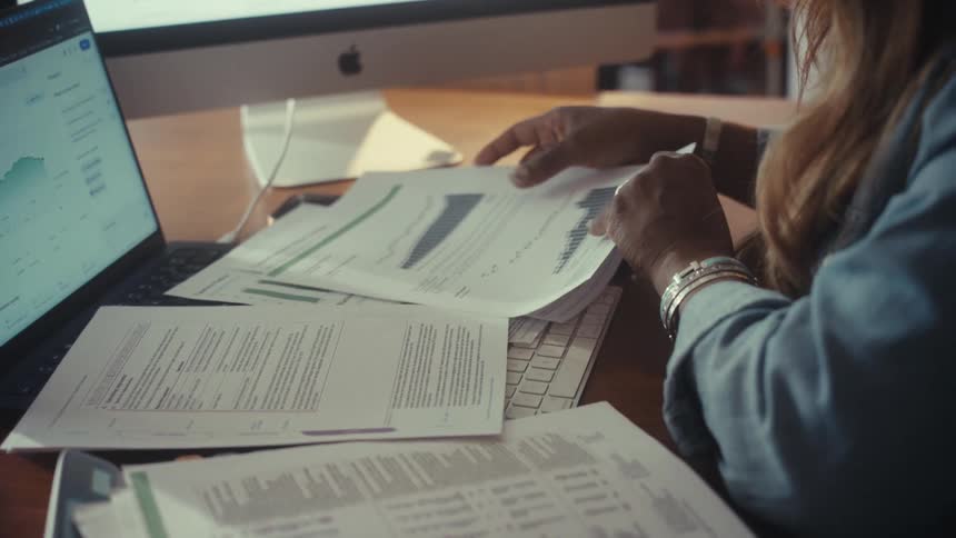 A financial advisor carefully reviews printed reports with charts and graphs at their desk in slow motion