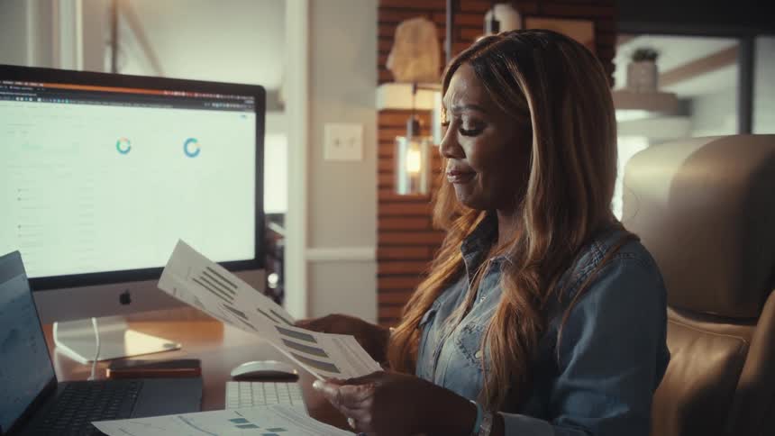 A professional African American woman meticulously reviews financial charts and graphs at her desk while working from her modern home office