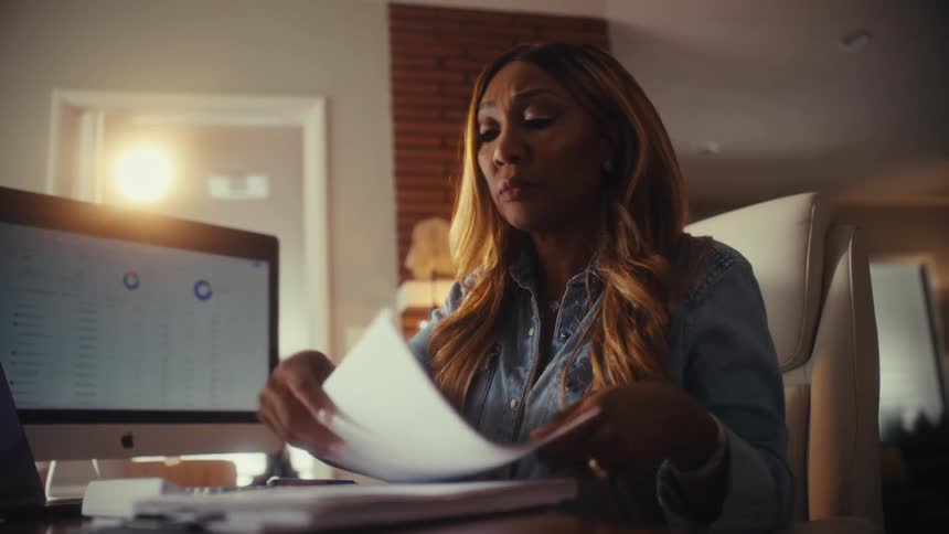 A focused businesswoman works late in her home office, analyzing financial charts and data on paperwork and her computer