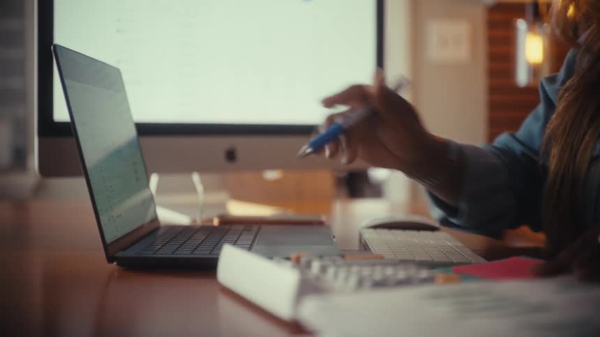 A woman works on her finances at her desk, using a calculator and referencing data on her laptop