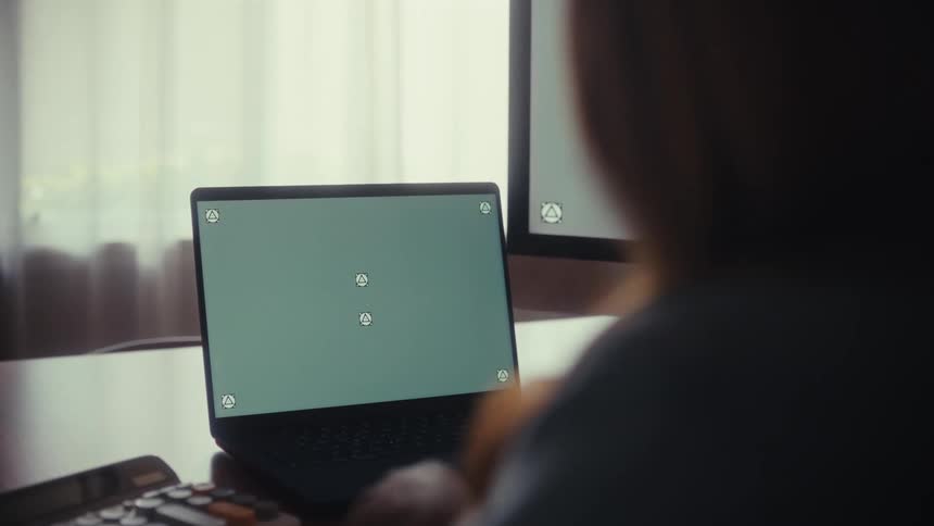 A person works at a desk with a laptop and desktop computer, both featuring green screens ready for custom content