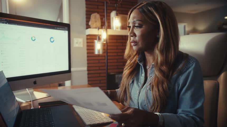 A concerned businesswoman analyzes financial data on multiple computers while cross-referencing paperwork in her modern home office