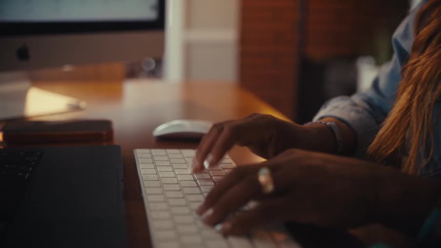 A close-up shot of a woman's hands typing and using a mouse while working at her computer in a warm home office setting
