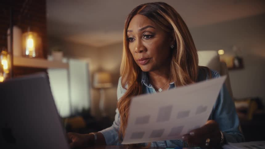A focused woman reviews a financial report and works on her laptop late at night in a warmly lit home office