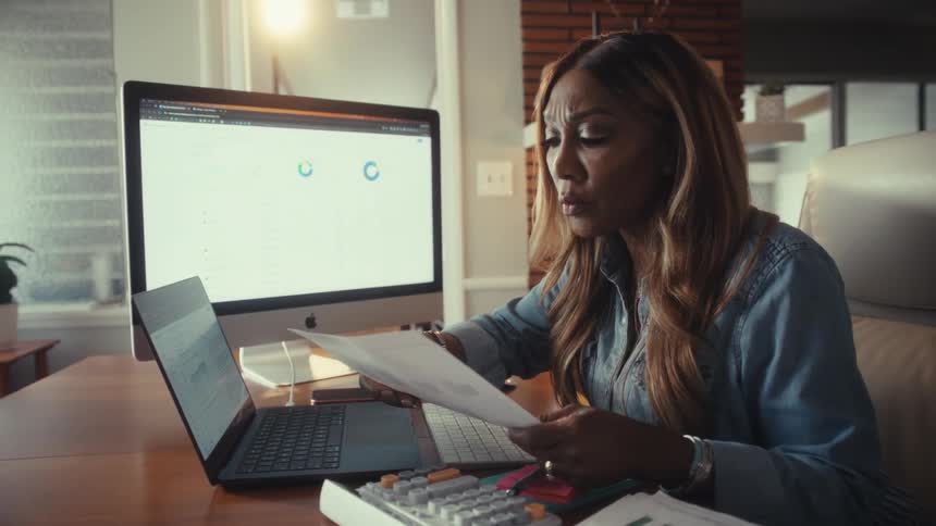 A focused businesswoman meticulously analyzes financial charts and documents on her computers while working from her modern home office