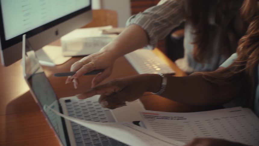 Close up on two businesswomen analyzing financial charts on a laptop while reviewing documents at a desk