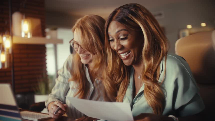 Two happy, diverse businesswomen laugh while collaborating on work together in a cozy, modern office
