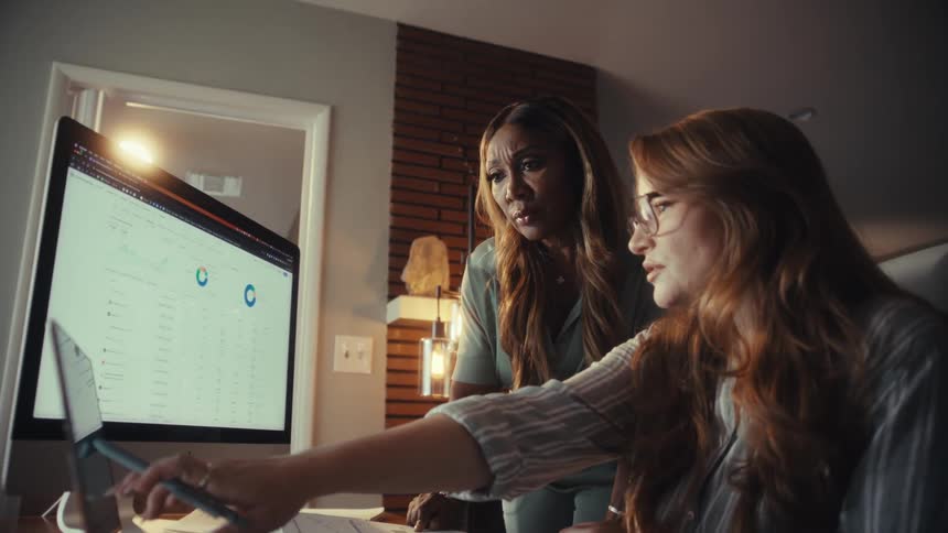 Two focused businesswomen collaborate, analyzing data and charts on computer screens in a low-lit office environment