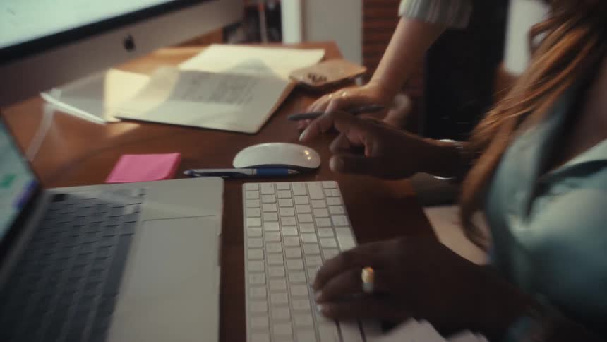 A close-up of two business professionals collaborating at a desk, with one typing on a keyboard while analyzing financial data on computer screens
